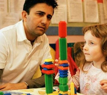 Male teacher and young girl play with coloured building bricks in the classroom.