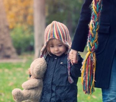 Child in wolly hat, carrying a teddy and holding the hand of an adult walking through a park.