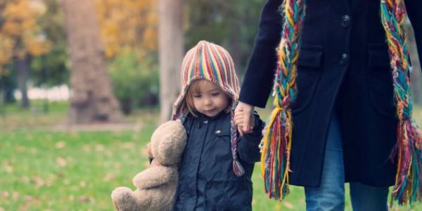 Child in wolly hat, carrying a teddy and holding the hand of an adult walking through a park.