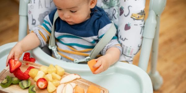 Baby in highchair plays with strawberries, kiwi and melon