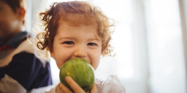 Little redheaded girl holds a green apple up in front of her face as she goes to take a bite. Happy eyes as she looks at the camera.