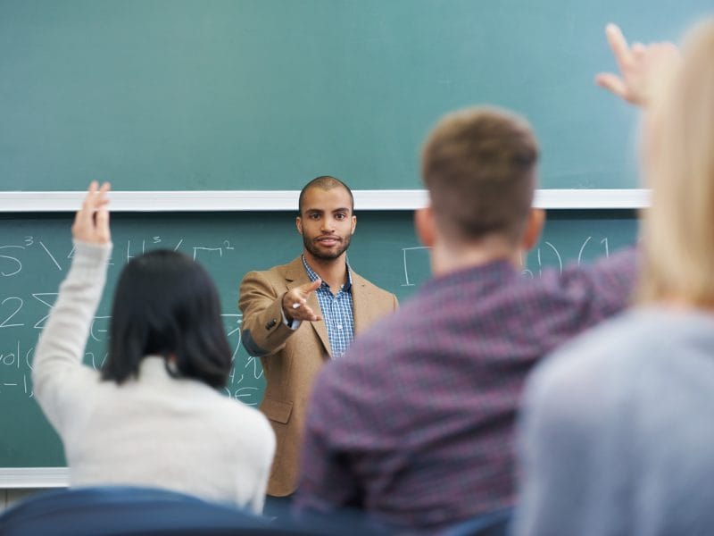 Young male teacher giving a lesson to his students. There are maths equations on the chalk board behind him and students have their hands up to answer his question.