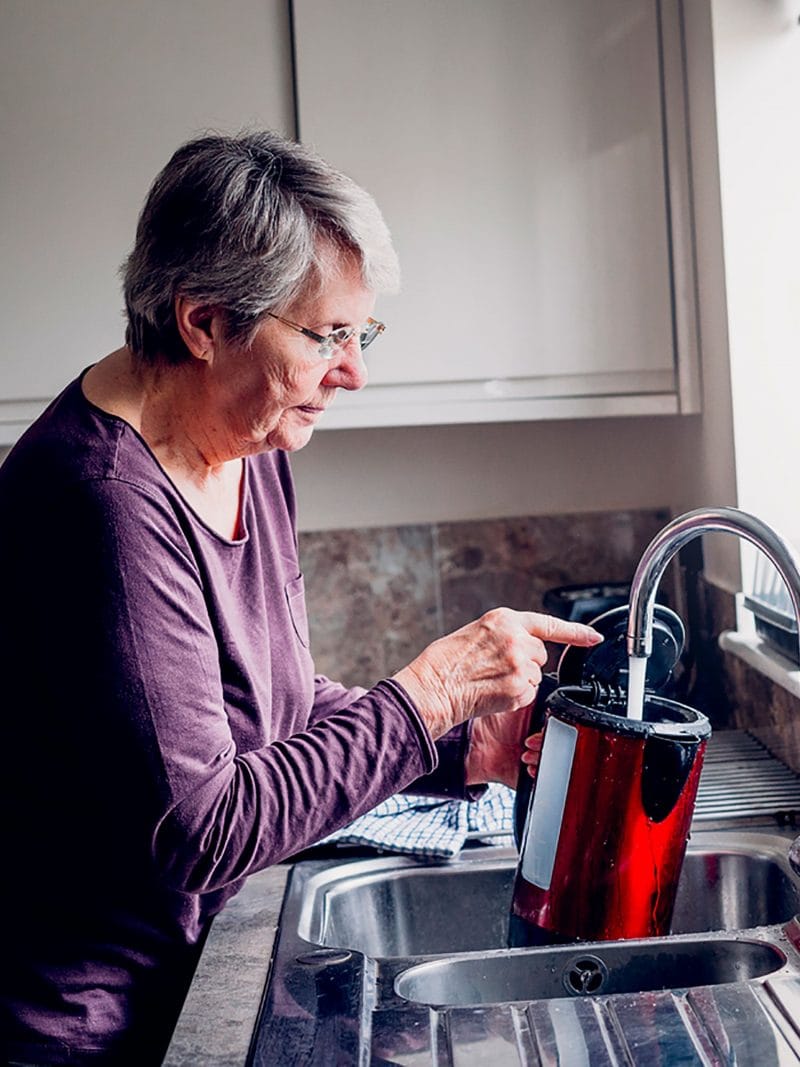 Older lady wearing purple top filling up kettle in kitchen sink by the window