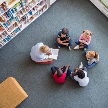 Top view of librarian sitting with five multiethnic children on floor. Teacher reading book to cute girls and young boys at school.