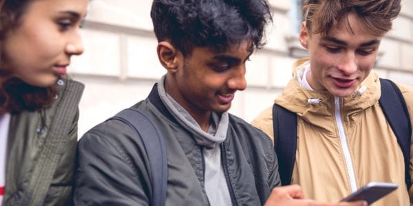 Three students wearing jackets smile and look at something on a mobile phone.