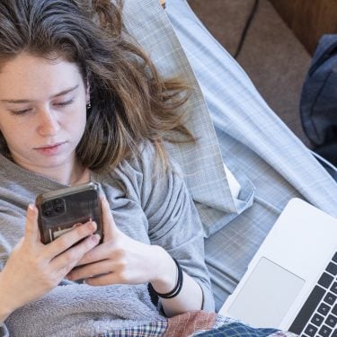 Teenage-girl-looks-at-smartphone-next-to-laptop-Growing-up-under-COVID-19-PROJ