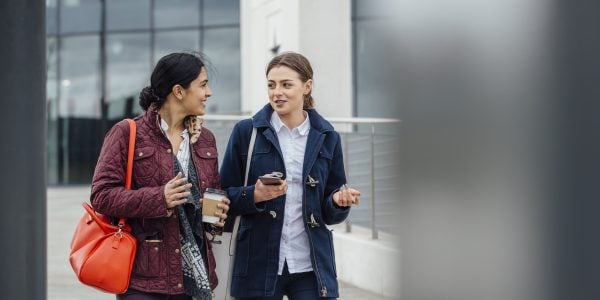 Businesswomen are walking to work together through the city. They are talking and one is holding a coffee while the other is holding a smartphone.