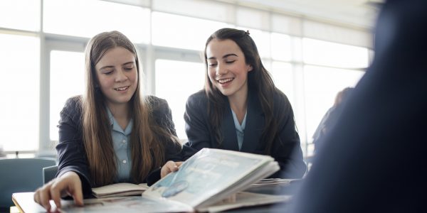 Teenager female students talking and looking through a text book during a lesson.