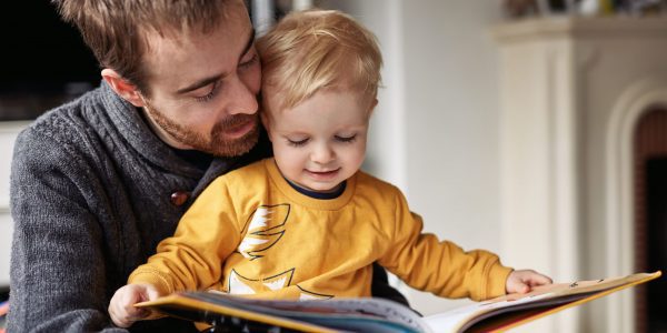 Young blonde boy wearing yellow jumper reads book with on father's lap.