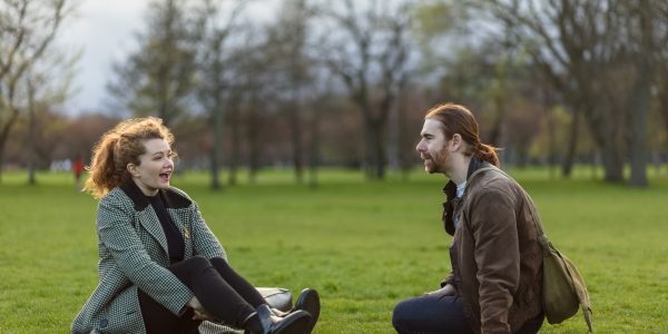 A caucasian man and woman, both with red hair are sitting on the grass and chatting in The Meadows, a park in Edinburgh, Scotland