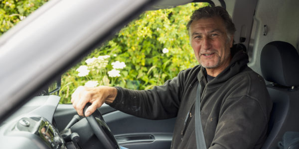 A shot of a senior, Caucasian man looking at the camera smiling while driving a van.