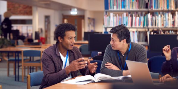 Teacher With Mature Male Adult Student Sitting At Table Working In College Library