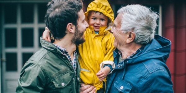 A father and grandfather hold up a little boy in a yellow rain jacket, all smiling