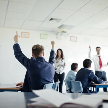 A classroom of children with their hands up to answer a question, with two teachers at the front of the classroom