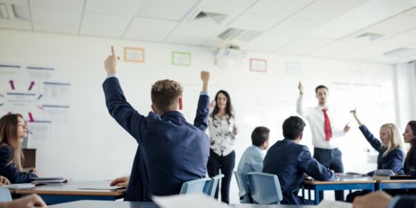 A classroom of children with their hands up to answer a question, with two teachers at the front of the classroom