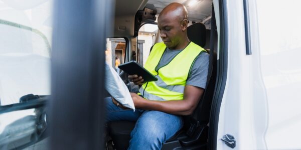 A man working as a delivery driver sits in the driver's seat, checking his list on a tablet