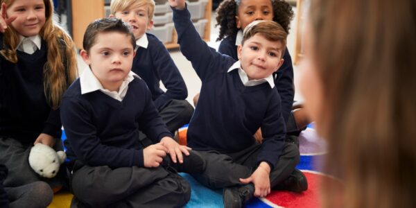 A group of primary school children sitting on the carpet listening to their teacher. One child is excitedly raising his hand to answer a question.