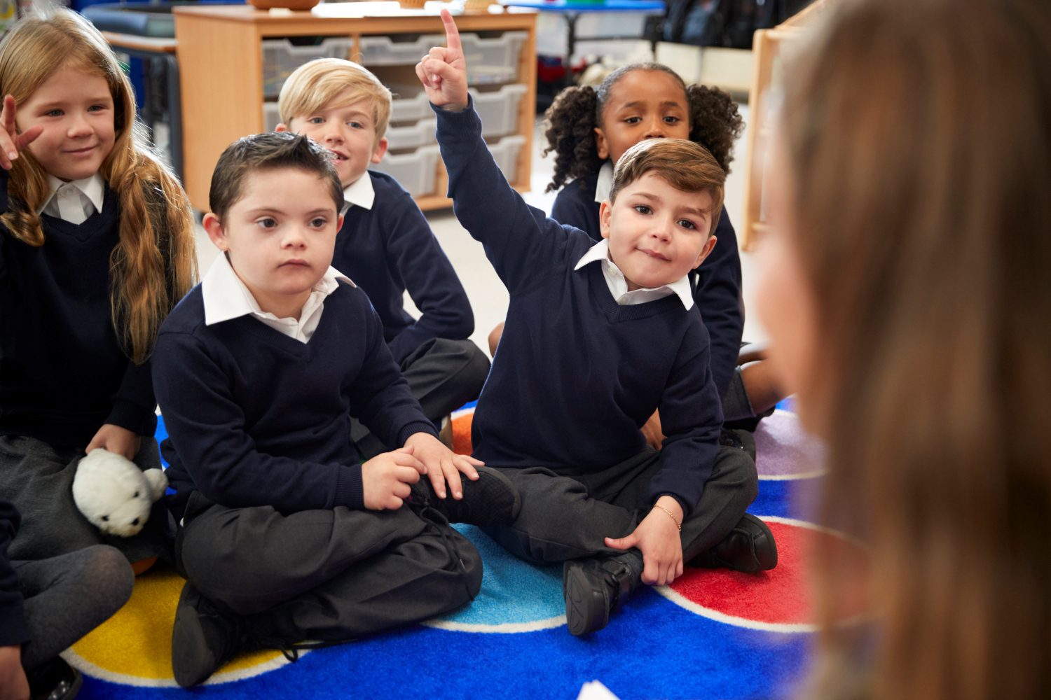 A group of primary school children sitting on the carpet listening to their teacher. One child is excitedly raising his hand to answer a question.