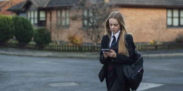 Girl in school uniform checks her phone while walking to secondary school, carrying a backpack