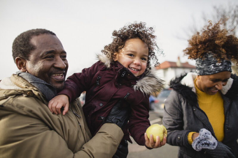 Grandparents having fun outdoors with their granddaughter, who is eating an apple and laughing.