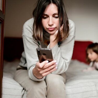 A woman is sat on the edge of a bed staring at her phone while her daughter is in the background.