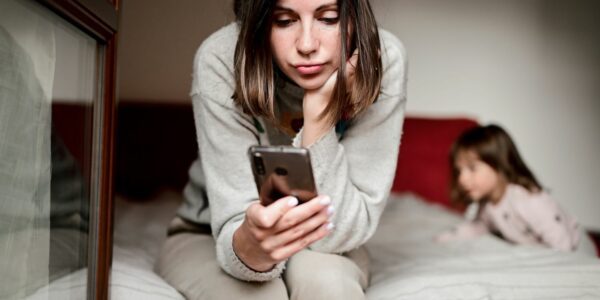 A woman is sat on the edge of a bed staring at her phone while her daughter is in the background.