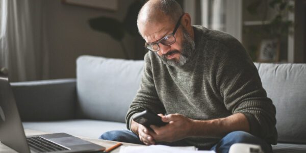 A middle-aged man sat at home is looking at this phone while sorting pension paperwork.