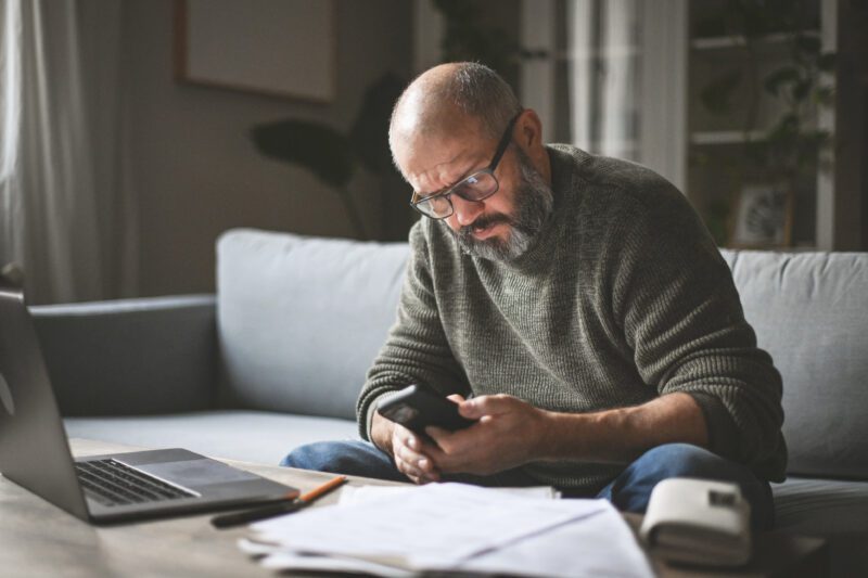 A middle-aged man sat at home is looking at this phone while sorting pension paperwork.