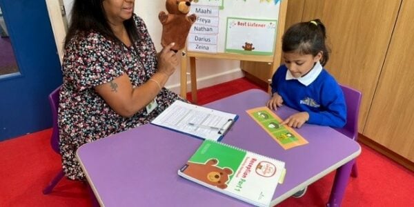 A teacher has a one on one session with a primary school student. The student is pointing at images and the teacher is holding a teddy bear puppet.