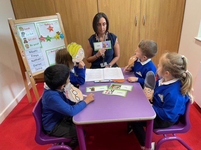 NELIatOxendon3 A teacher sits at a table with 4 primary school students. They are looking and talking about images and words on cards.