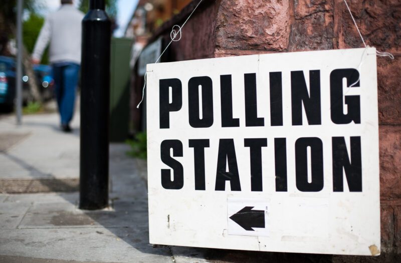 A sign to a polling station hangs off a brick wall. In the background a man is walking in the direction of the sign.
