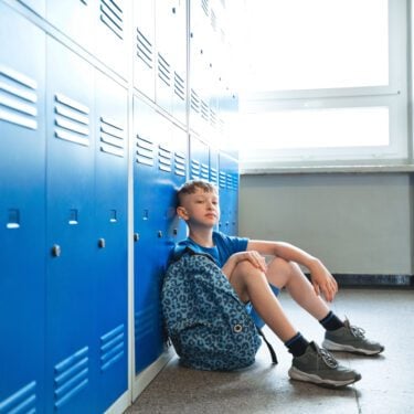 A boy, wearing his PE kit, leans against the lockers. He's on his own in a corridor at school and is looking at the camera.