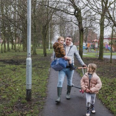 A front view of a father and his two children. He is carrying his young son in his arms and his daughter is riding her scooter along the footpath as he pushes his son's scooter along the way.