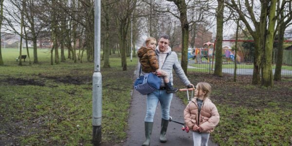 A front view of a father and his two children. He is carrying his young son in his arms and his daughter is riding her scooter along the footpath as he pushes his son's scooter along the way.