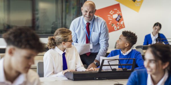 Secondary school music teacher smiles at students working together at a keyboard.