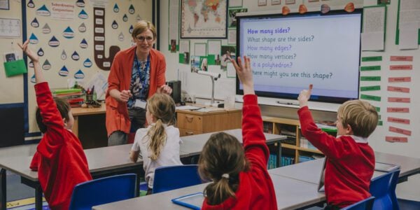 Female teacher is teaching shapes to her primary school students. She is asking hem a question and some of the students have their hand in the air to answer.