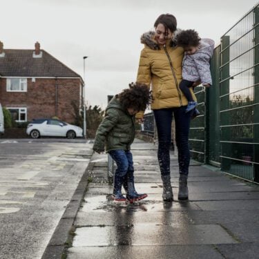 A young child splashing in a puddle next to his guardian and sister.