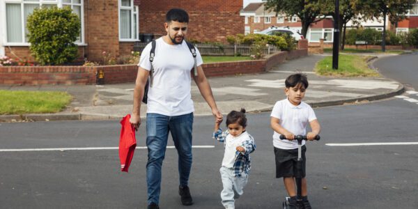 Father taking his children to school. He's carrying his child's book bag. The older child is riding a scooter to school, and the younger child is walking holding their dad's hand.