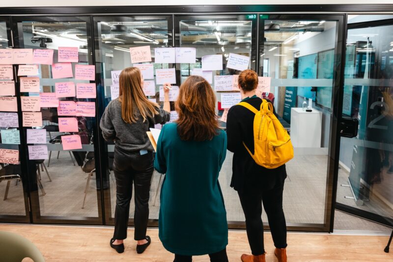 Three women stand looking at a wall of post-it notes in the Nuffield Foundation office. One of them is writing on a post-it.
