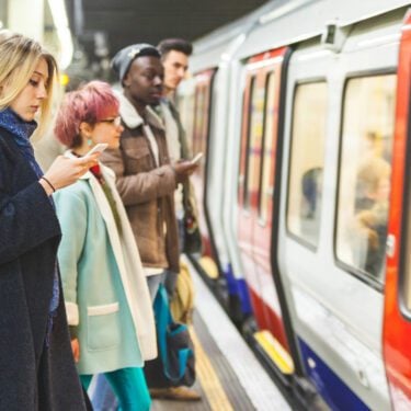 Commuters waiting to get on the tube. There are two men and two women. One woman is looking at her phone, looking concerned.