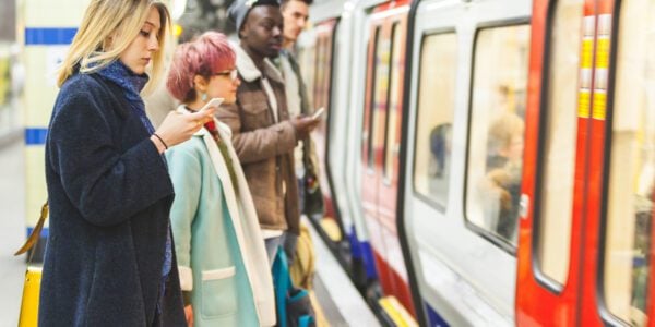 Commuters waiting to get on the tube. There are two men and two women. One woman is looking at her phone, looking concerned.