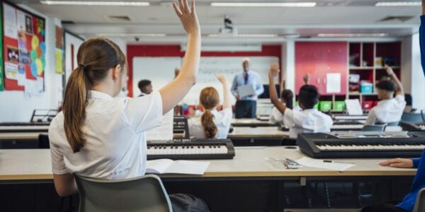 A wide-view shot of teenage music students in a classroom learning how to play the keyboard with their teacher at a school. The students are wearing school uniforms and the teacher is smartly dressed.