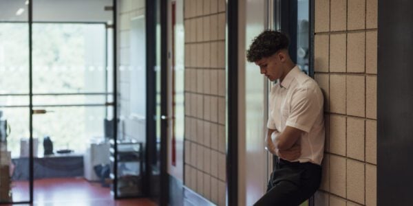 A side view of a young teenage boy who is looking forlorn and sad after being sent out of the classroom. He has his arms folded and is looking down.