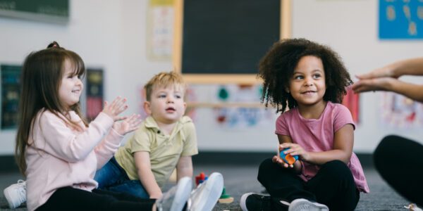 A group of toddler students sit together on the floor of their preschool classroom, playing with blocks, laughing and smiling with each other.