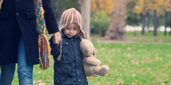young girl walking through park hugging a teddy bear and holding-her mums hand
