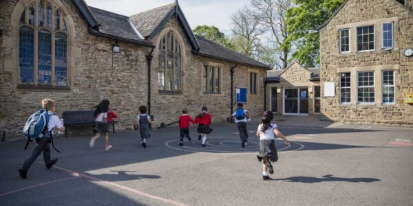 Rear view of children running in their school yard. They are all running towards the door with their backpacks on.