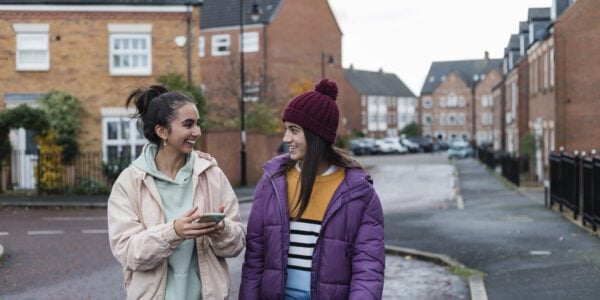 Two teenage sisters spending time together outdoors, laughing and talking on a walk, using a mobile phone together.