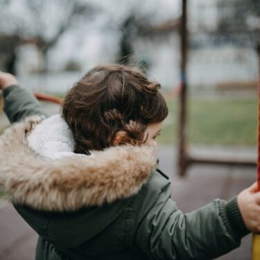 Rear View Of Girl Holding Ropes At Playground. She is wearing a coat and the sky is grey.