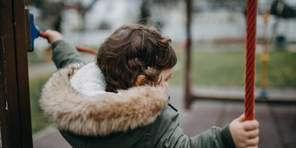 Rear View Of Girl Holding Ropes At Playground. She is wearing a coat and the sky is grey.