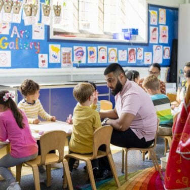 Pre-school students sitting in an art classroom being taught by a teacher. The classroom is colourful and the children are sat at a big table.